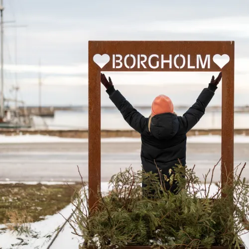 En glad tjej står med armarna uppåt vid Borgholms skylt i gästhamnen.