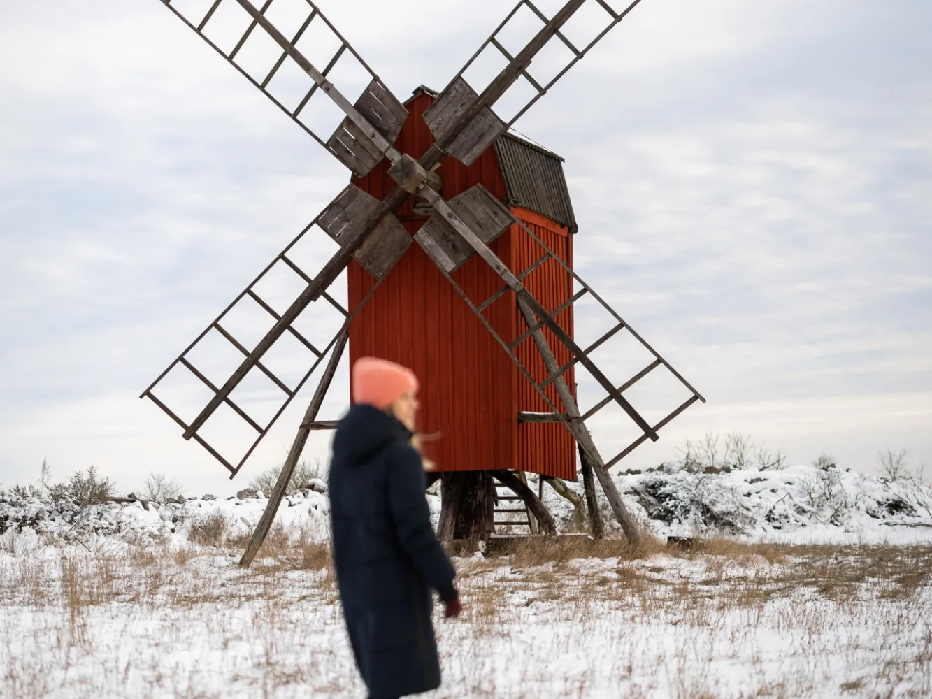 A woman stands in a winter setting in front of a red Öland windmill.