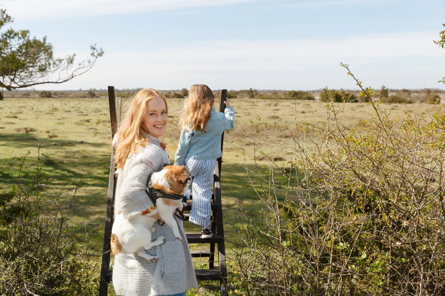 En kvinna håller en hund medan ett barn klättrar över en grind i ett öppet landskap.
