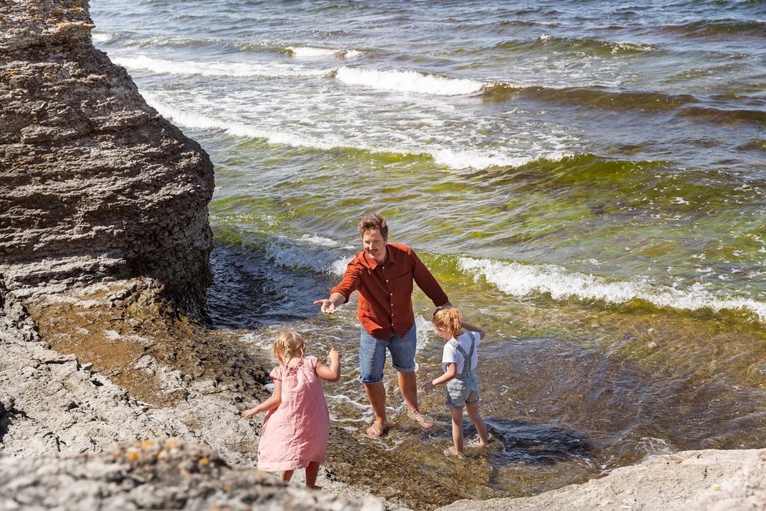 En vuxen och två barn leker vid strandkanten mellan raukarna
