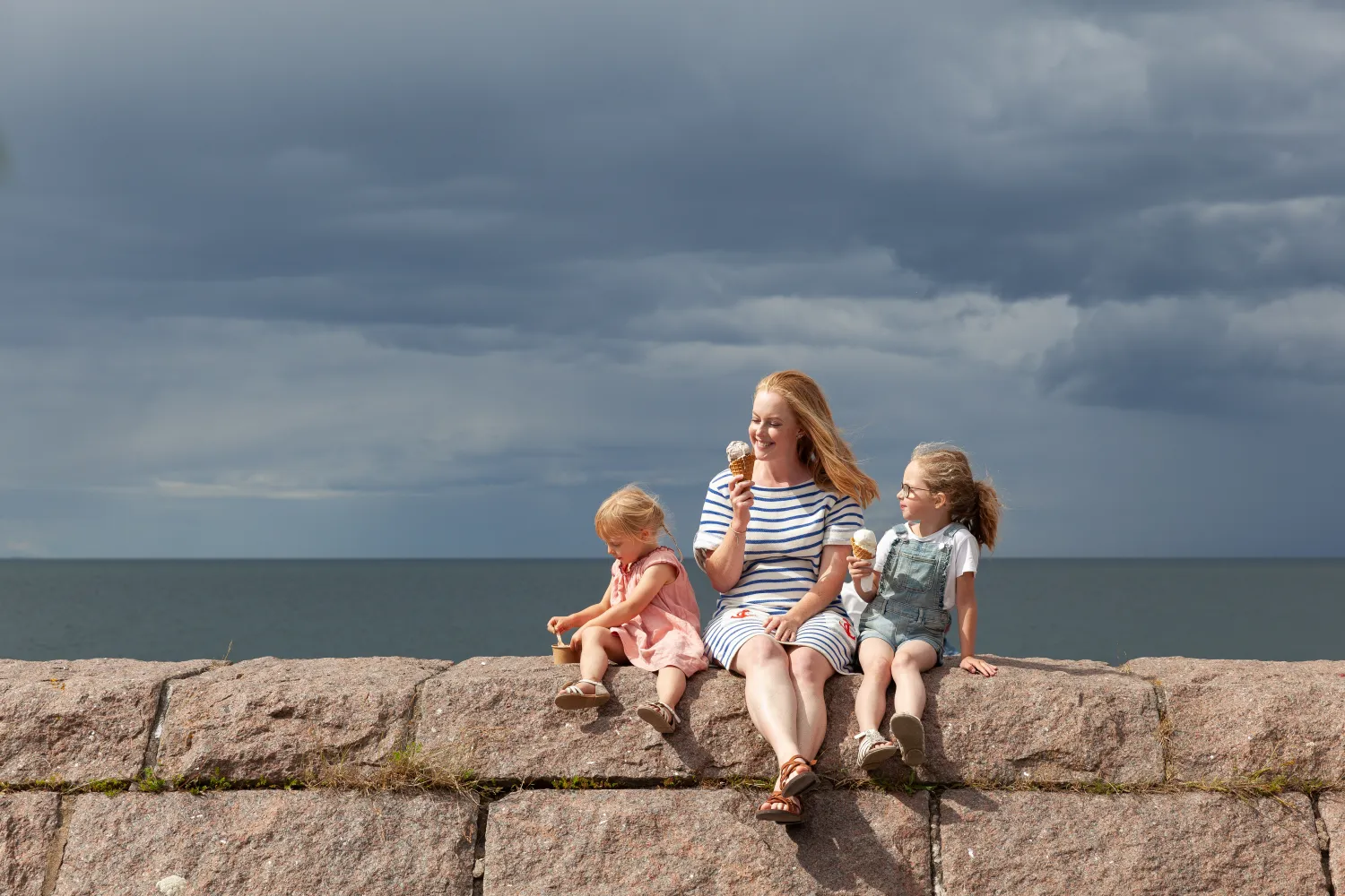 En kvinna och två barn sitter och äter glass på en stenmur