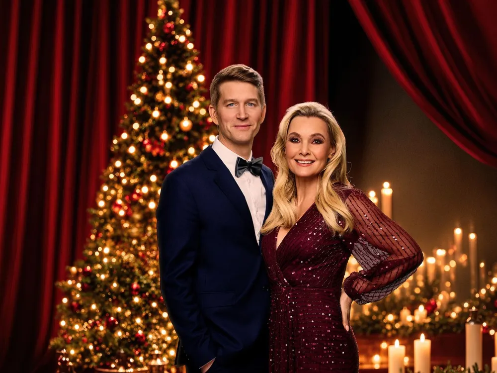 Ett elegant par poserar framför en upplyst julgran med stjärna, röda draperier och tända ljus i bakgrunden.