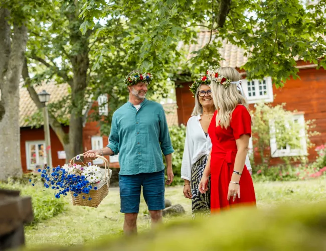 En man med midsommarkrans i håret håller i korg full av midsommarblommor och ler glatt mot två kvinnor i varsin midsommarkrans.