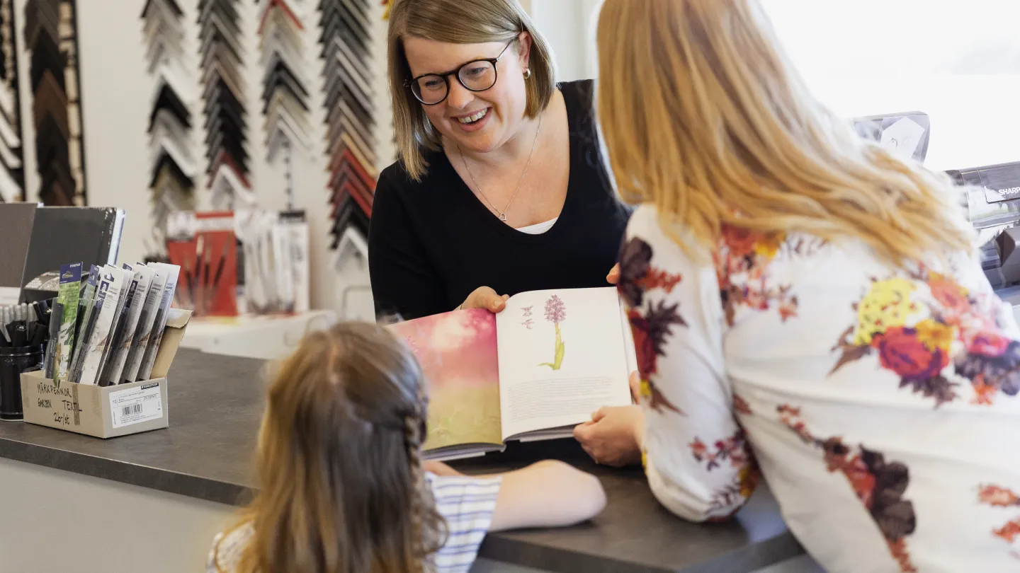 En kvinna visar en bok om orkidéer för en annan kvinna och ett barn i en butik.