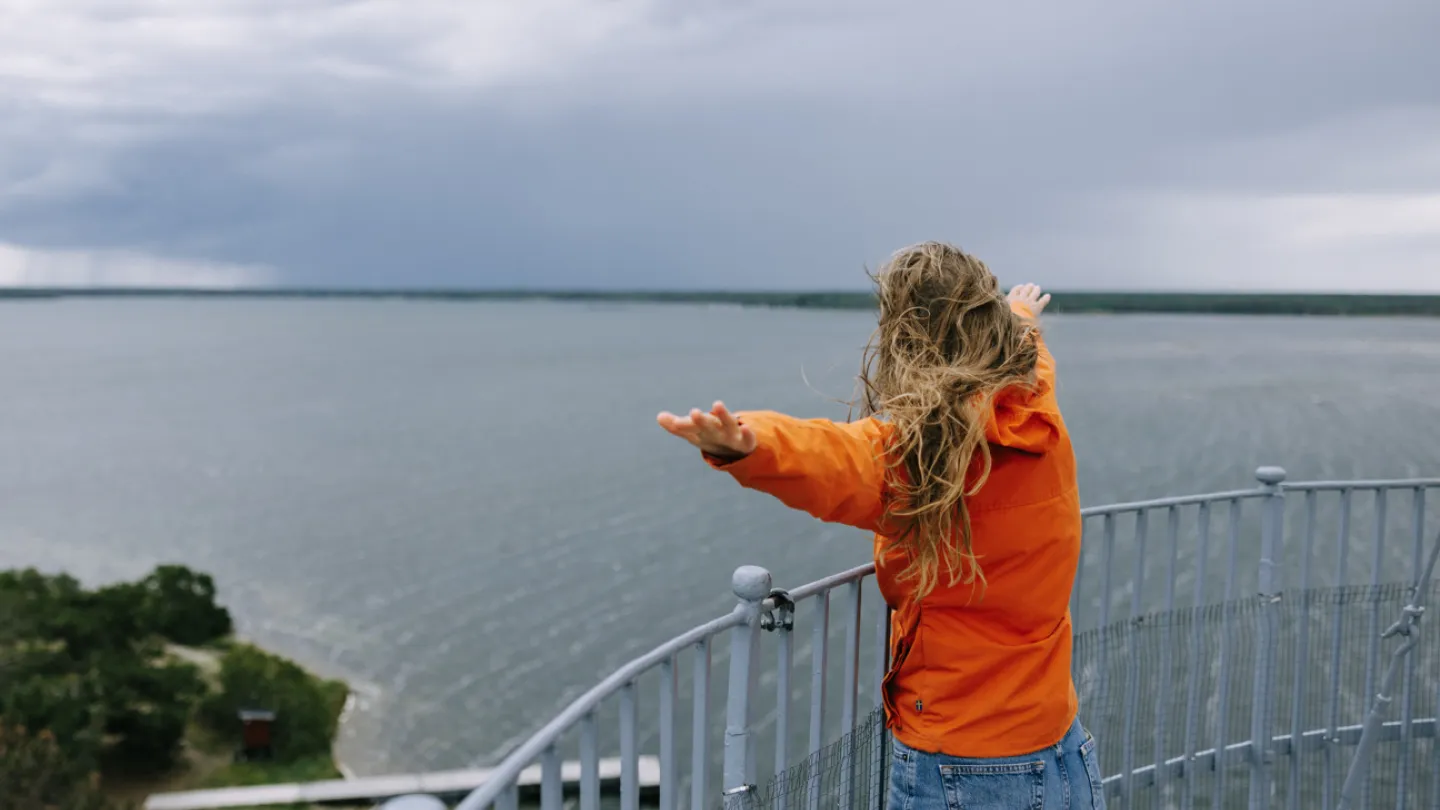 En tjej i röd jacka står med armarna utåt och blickar ut mot havet.