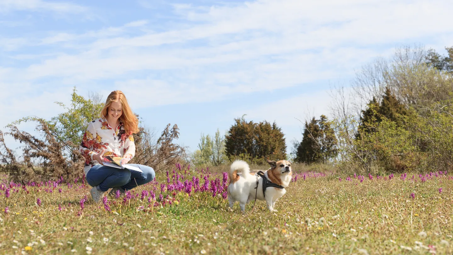 n kvinna sitter i en äng och läser i en bok om orkidéer, bredvid står en liten hund 