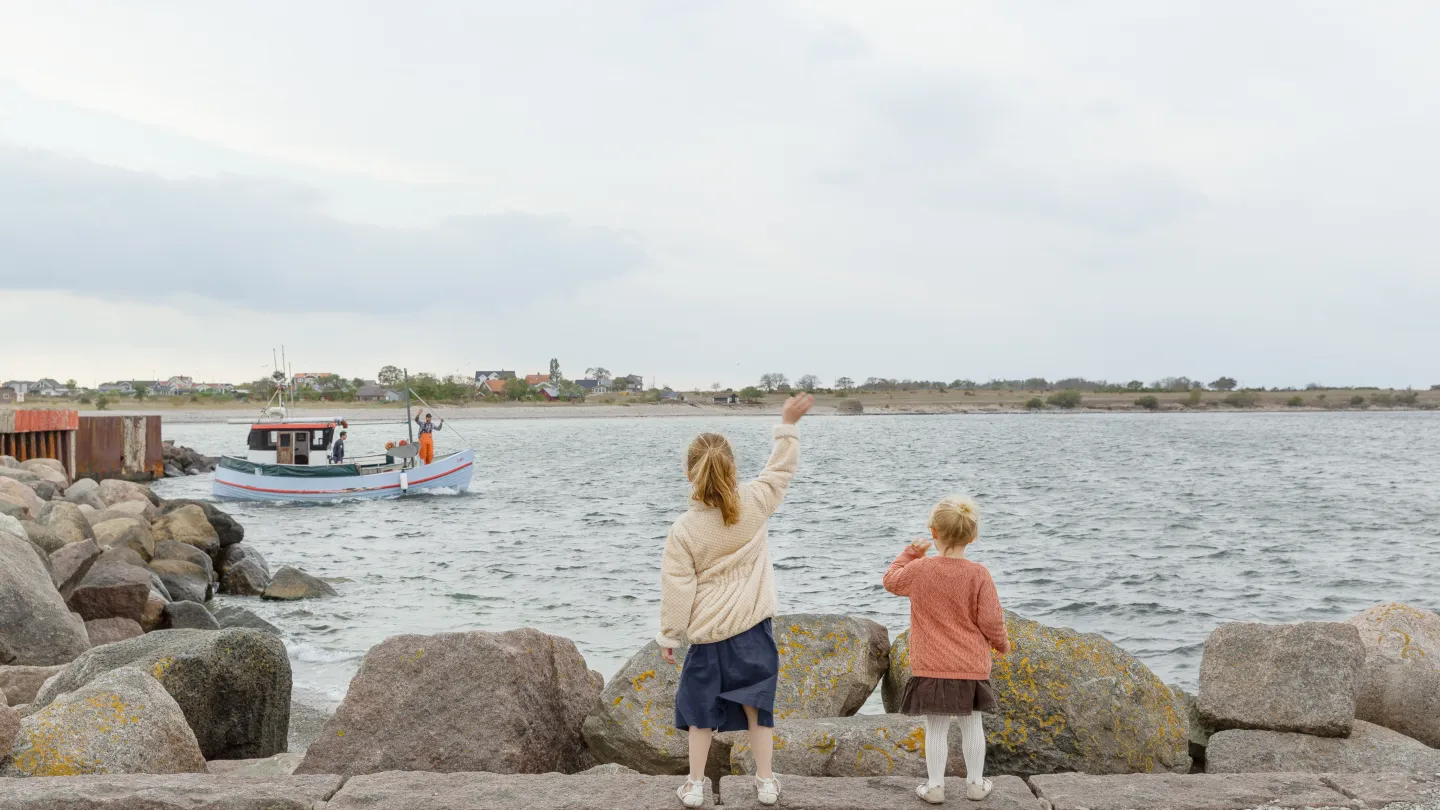 Två barn står vid vattnet och vinkar mot en båt.