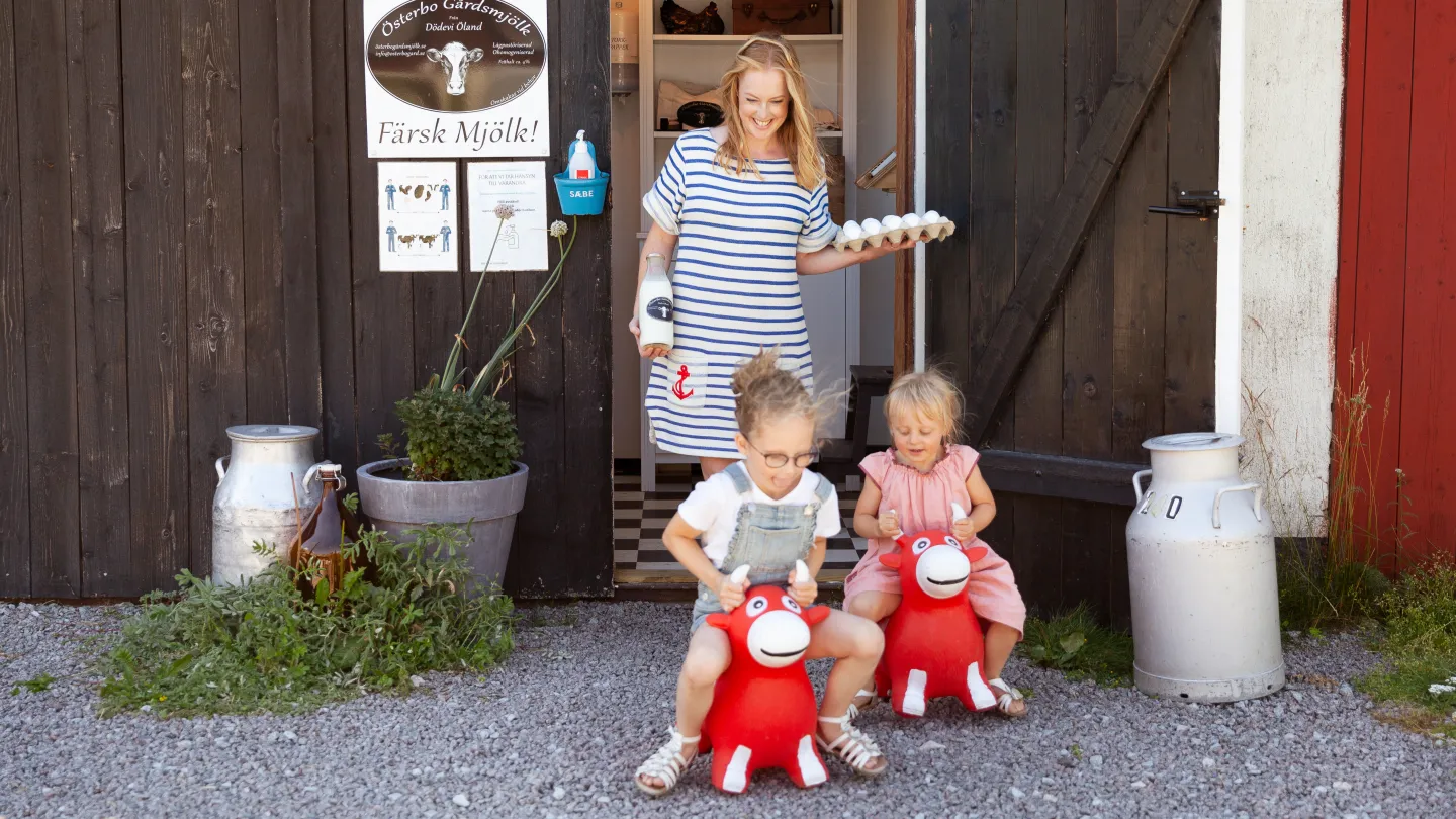 n kvinna och två barn utanför en gårdsbutik med färsk mjölk och ägg.