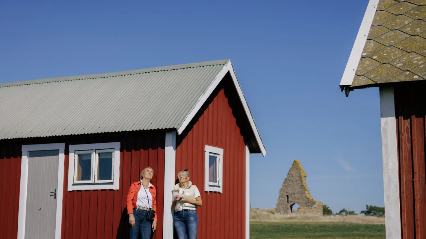 Två damer står mot en husvägg, blundar och njuter av vårsolen vid Kapelludden fyr på Öland.